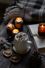 Autumn cozy home composition with hot chocolate with marshmallow and candles. Aromatherapy on a grey fall morning, atmosphere of cosiness and relax. Wooden background, books, close up.
