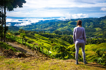 girl stands on hill admiring idyllic landscape of Costa Rican mountains; green slopes bathed in...