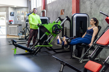 Fototapeta na wymiar Personal trainer coaching a young caucasian woman, legs exercises with machine in the gym, workout concept