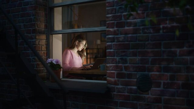 Young Woman with Blond Hair Working from Home on Laptop Computer in Cosy Loft Brooklyn Style Apartment. Female Smiling, Checking Social Media, Browsing Internet. View Through the Window From Outside