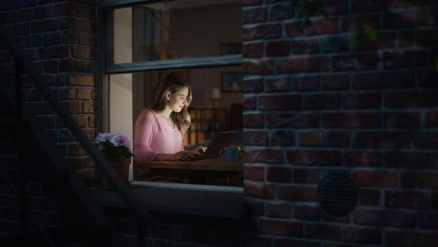 Beautiful Blond Woman Works on a Laptop while Sitting at Her Desk in the Bedroom. Female Professional Freelancer Working from Cozy Home at Night. View Through the Window From Outside