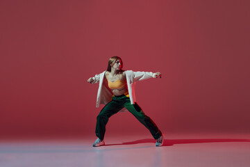 Dynamic portrait of young girl dancing hip-hop or contemporary dance isolated on dark red background. Experimental dance. Music, dance, active lifestyle, fashion, style