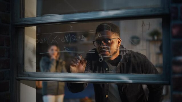African American Young Man Using an Erasable Marker to Solve a Math Equation on Window Glass. Black Male Tutor Writing a Physics Theory and Explaining it to Fellow Female Student.