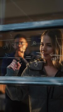 Vertical Shot: Caucasian Young Woman Using an Erasable Marker to Solve a Math Equation on Window Glass. White Female Tutor Writing a Physics Theory and Explaining it to Fellow male Student.