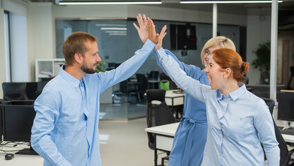 Obraz premium Colleagues give a high five in the office. Red-haired woman, blonde and bearded man in a denim shirt in the office.