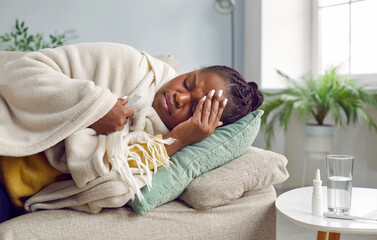 Sick young african american woman is not feeling well and is lying at home on the couch with her...