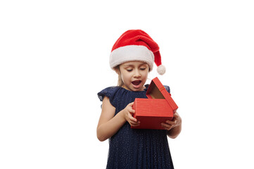 Surprised little child girl in Santa hat and elegant blue dress expressing excitement while unpacking a red gift box with cute Christmas present, isolated over white background. Time to open presents