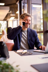 Mature Businessman Working On Laptop At Desk In Office Talking On Mobile Phone