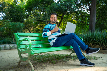 Fototapeta premium Young indian man using laptop and showing thumps up at park.