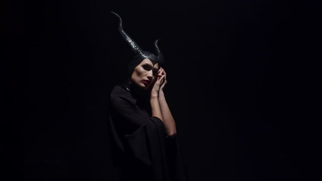 Portrait of an evil witch villain woman from a fairy tale with horns is standing, touching her face with her hands, posing in a dark black studio