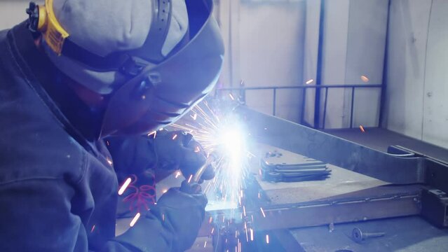 close-up shot of young professionals welder wearing uniform, protective helmet and gloves is working on welding metal frame arc with sparks flying off it in her workshop