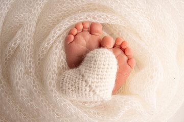 The tiny foot of a newborn baby. Soft feet of a new born in a white wool blanket. Close up of toes, heels and feet of a newborn. Knitted white heart in the legs of a baby. Macro photography. - obrazy, fototapety, plakaty