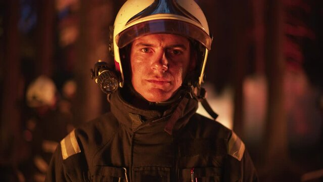Portrait of a Brave Handsome Young Adult Firefighter in Safety Uniform and a Helmet with a Flashlight. Professional Fireman Looking at Camera. Wildland Fire in a Forest in the Background.