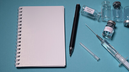 Blank notebook with pen on a blue background. Empty vaccine vials and a syringe with a needle lie on the table. Concept: Vaccine, injection, money, counting.
