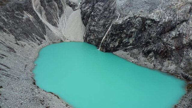 Drone flyover of Laguna 69 (lake 69) or Laguna Sesentinueve, a luminescent green turquoise chemical lake in Peru. Camera flies forwards low over the lake with the glacial waterfall in the background.