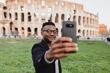 an african on vacation takes a selfie in front of the colosseum smiling.