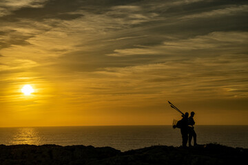 Czarna sylwetka rybaków stojących na klifie, pomarańczowy zachód słońca, przylądek Świętego Wincentego (port. Cabo de São Vicente) Portugalia - obrazy, fototapety, plakaty