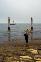 Fragment nabrzeża rzeki Tag w Portugalii ze słynnymi kolumnami (port. Cais das Colunas) w pochmurny, deszczowy dzień. Sylwetka kobiety z kolorowym parasolem. © Aneta