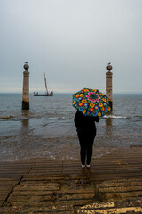 Fragment nabrzeża rzeki Tag w Portugalii ze słynnymi kolumnami (port. Cais das Colunas) w pochmurny, deszczowy dzień. Sylwetka kobiety z kolorowym parasolem. © Aneta