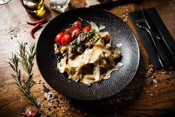 Black Angus pappardelle pasta with beef, champignon and parmegano cheese on a black plate. Delicious healthy mediterranean traditional food served on a table for lunch in modern gourmet restaurant  - 552609380