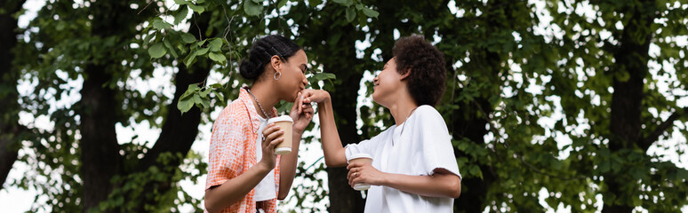 Naklejka premium joyful african american lesbian woman holding coffee to go and kissing hand of girlfriend in park, banner.