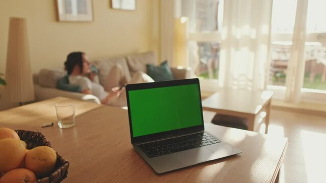 Foreground on the table is laptop with green screen chroma key, in the background girl is sitting, soft focus