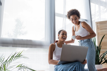 young african american lesbian woman using laptop near joyful girlfriend holding cup of coffee.