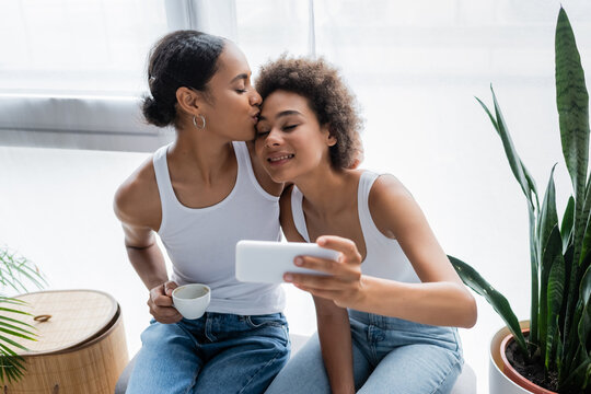 cheerful african american lesbian woman taking selfie while girlfriend holding cup of coffee and kissing her head.