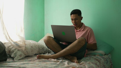 South American hispanic young man using laptop in bedroom. A Brazilian latin person browsing internet online sitting in bed at home looking at computer screen
