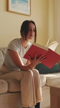 VERTICAL VIDEO, Young woman reading book sitting on the sofa at home