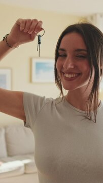 VERTICAL VIDEO, Young beautiful woman raising apartment keys in her hand and smiling at camera while standing at home