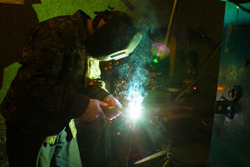 A masked worker performs spot welding work in his workshop, clamping a workpiece in a vise on a workbench