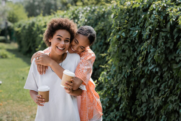 happy african american lesbian couple holding paper cups with coffee to go while hugging in park.