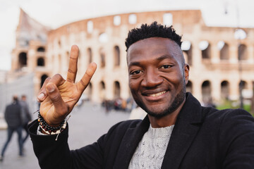 a very smiling african boy on holiday at the colosseum takes a selfie showing two hand fingers.