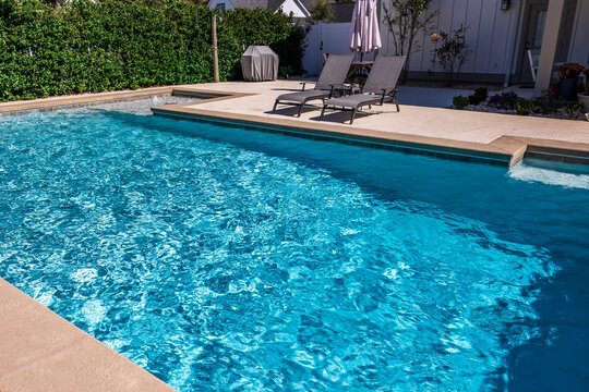 A rectangular new swimming pool with tan concrete edges in the fenced backyard of a new construction house