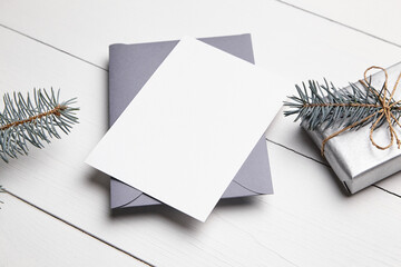 Christmas greeting card mockup with grey envelope, silver color gift box and green fir tree branch on white wooden table. Empty winter holiday card