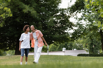 full length of joyful african american lesbian woman hugging happy girlfriend and walking in green park.