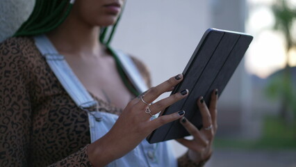 One young black woman using tablet outdoors. African American adult girl standing outside leaning on wall holding device touching screen