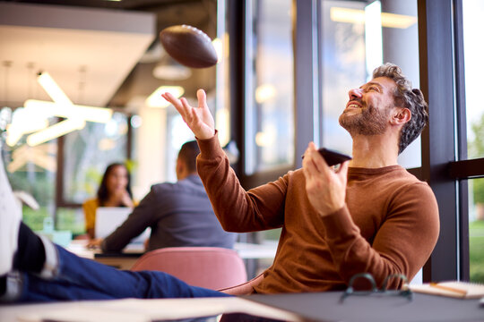 Mature Businessman With Feet On Desk In Office Talking Into Mic Of Mobile Phone Catching Football