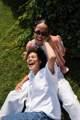 happy african american lesbian woman hugging cheerful girlfriend in sunglasses while sitting on lawn.