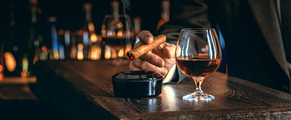 Man's hands with a cigar, elegant glass of brandy on the bar counter. Alcoholic drinks, cognac, whiskey, port, brandy, rum, scotch, bourbon. Vintage wooden table in a pub at night - obrazy, fototapety, plakaty