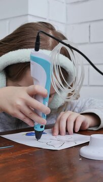 A cute European girl in fluffy headphones draws a picture with a 3D pen. The child is careful and attentive in using the electronic 3D pencil. Dolly shot vertical video