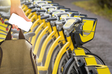 Close-up of a hand with a smartphone and a backpack in front of a row of rental bikes. Blank screen, you can paste your content