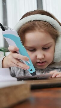 cute european girl in fluffy headphones draws with a turquoise 3d pen at a brown wooden table. After the end of the lesson, the girl puts the pen in the stand in the foreground. Contemporary art