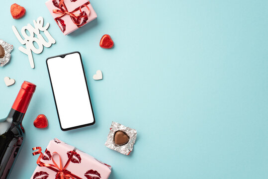 Saint Valentine's Day concept. Top view photo of smartphone wine bottle present boxes heart shaped candies and inscription love you on isolated pastel blue background with copyspace