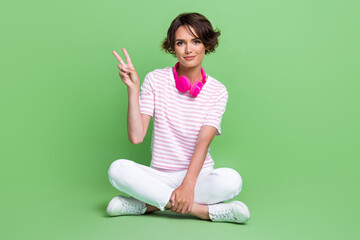Full size photo of pretty young lady sit folded legs earphones show v-sign dressed trendy striped...
