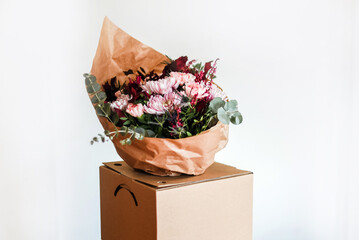 Un ramo de flores en tonos rosas y granates envuelto en papel marrón dentro de una caja de...