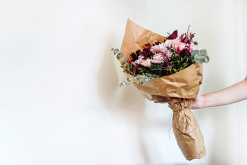 Una mano sujetando un ramo de flores en tonos rosas y granates envuelto en papel marrón. Eucalipto...