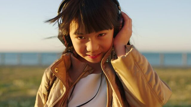 Face, music headphones and Asian kid at beach park streaming radio or podcast on holiday. Vacation, relax and happy girl from Japan listening to audio, song or sound track with head dance outdoors.