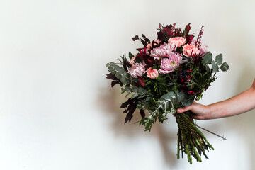 Una mano sujetando un ramo de flores en tonos rosas y granates. Eucalipto plateado, crisantemo, clavel, peonía y cardo azul.